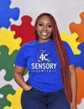 Woman wearing the royal blue 4C Sensory Center tee with dark jeans, in front of a colorful puzzle-piece backdrop